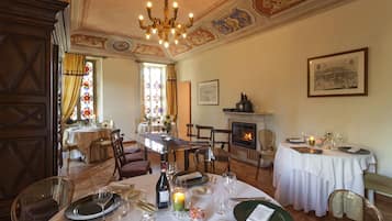 Uma área de refeições com mesas redondas, cadeiras e um candelabro. Há uma lareira e uma janela de vitrais ao fundo.