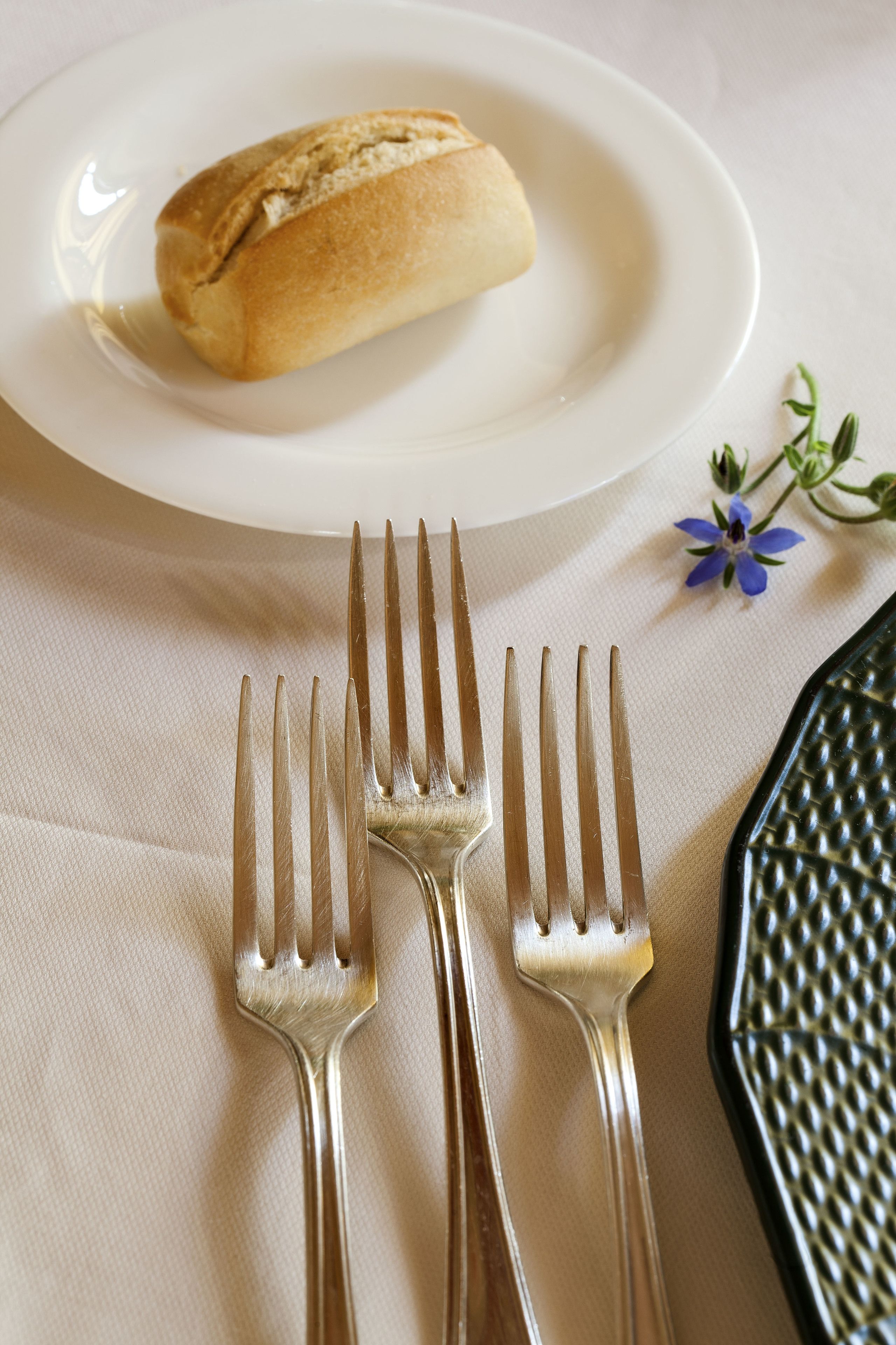 A white plate with a loaf of bread, three forks, and a small blue flower on a beige tablecloth.