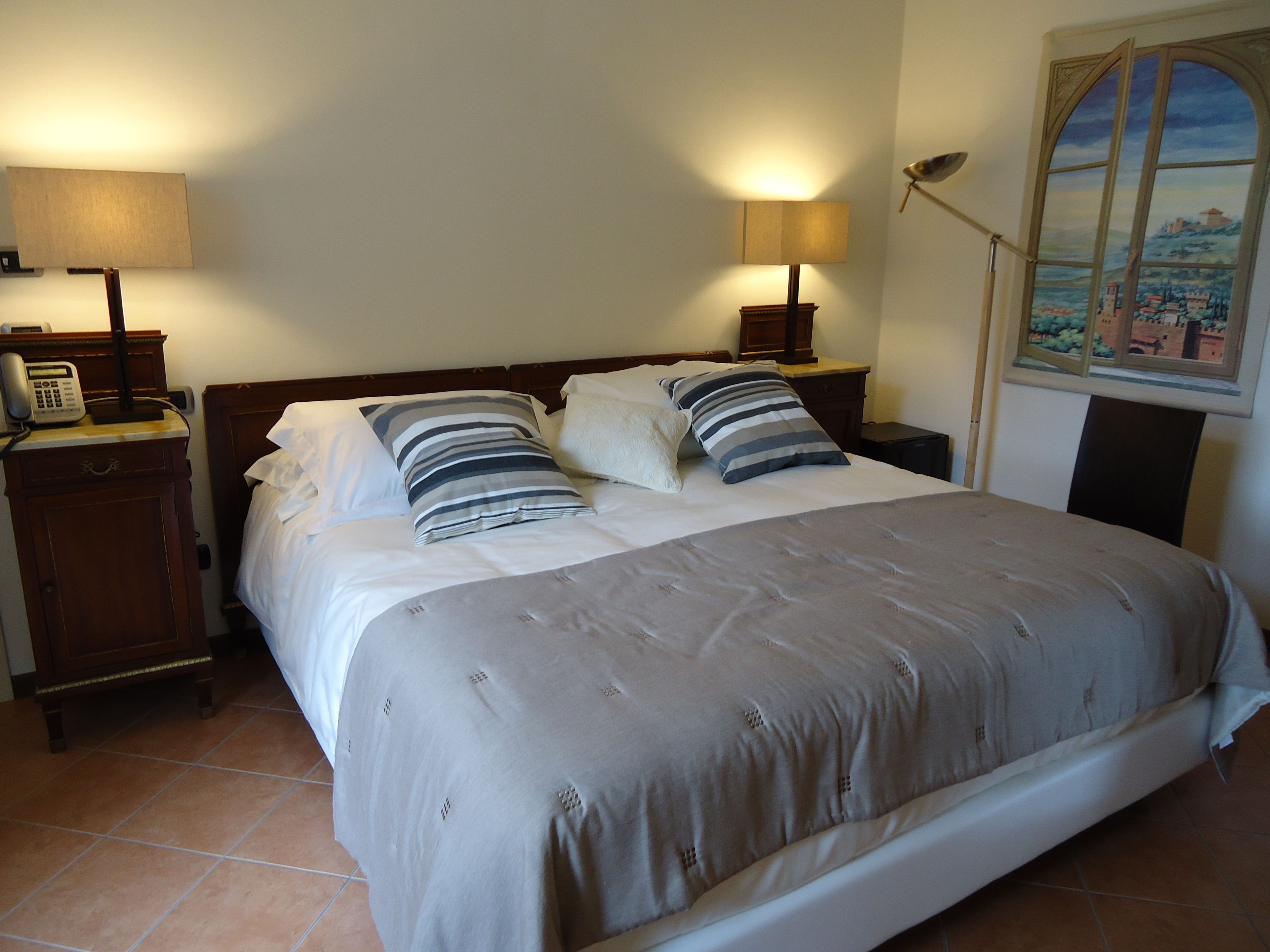 Uma cama bem arrumada com colcha cinza e almofadas listradas, duas luminárias de cabeceira, um telefone e um quadro de uma paisagem na parede.