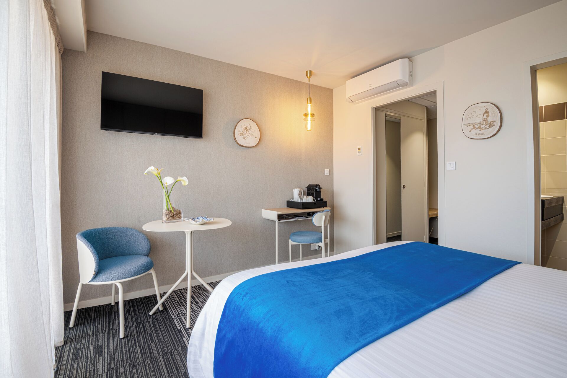 A hotel room with a bed, a desk, a chair, a small table with a vase of flowers, a flat-screen TV mounted on the wall, and a door leading to a bathroom.