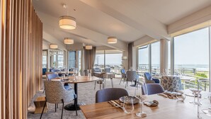 Un comedor moderno con techos altos, ventanas amplias y vista al océano. Las mesas están dispuestas con copas de vino y platos, y hay diversas opciones de asientos, incluyendo sillas y sofás.