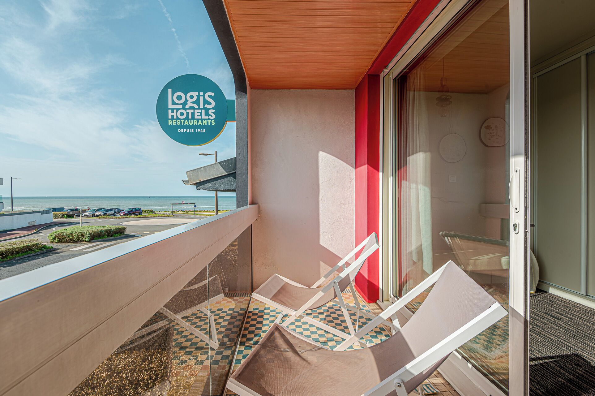 A balcony with two lounge chairs, a red wall, and a view of the sea.