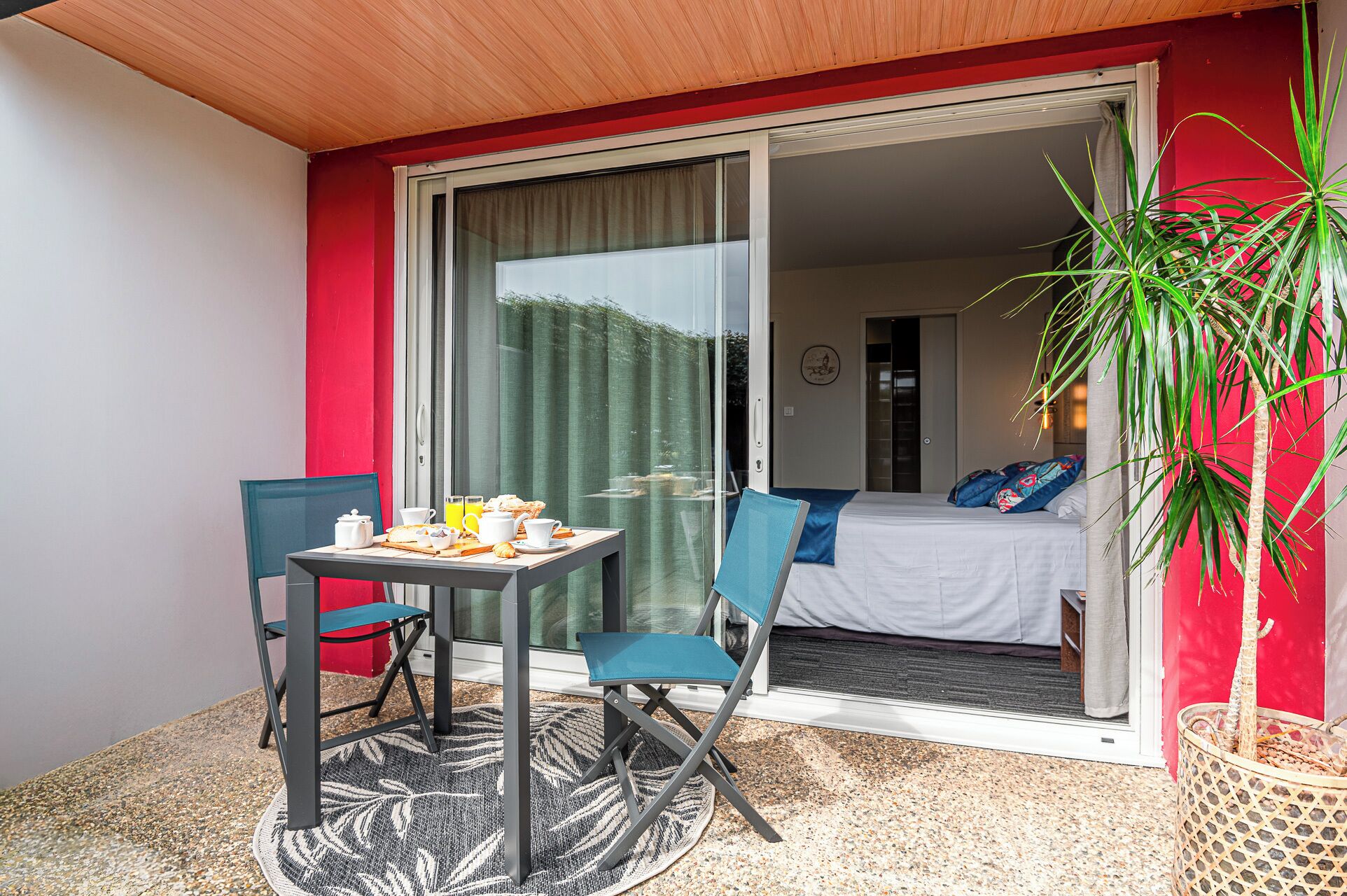 Un balcón con una mesa dispuesta para el desayuno, una silla y una cama visible a través de una puerta corrediza abierta.