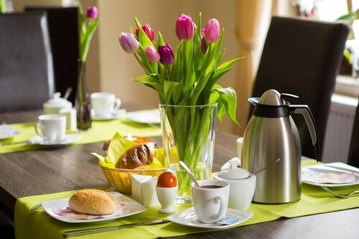 A breakfast setting with a coffee pot, cups, a plate with a pastry, a glass vase with tulips, and a basket of bread.