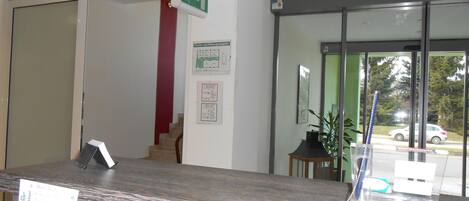 A reception desk with a computer monitor, a plant, and a window with a view of a street and parked cars.