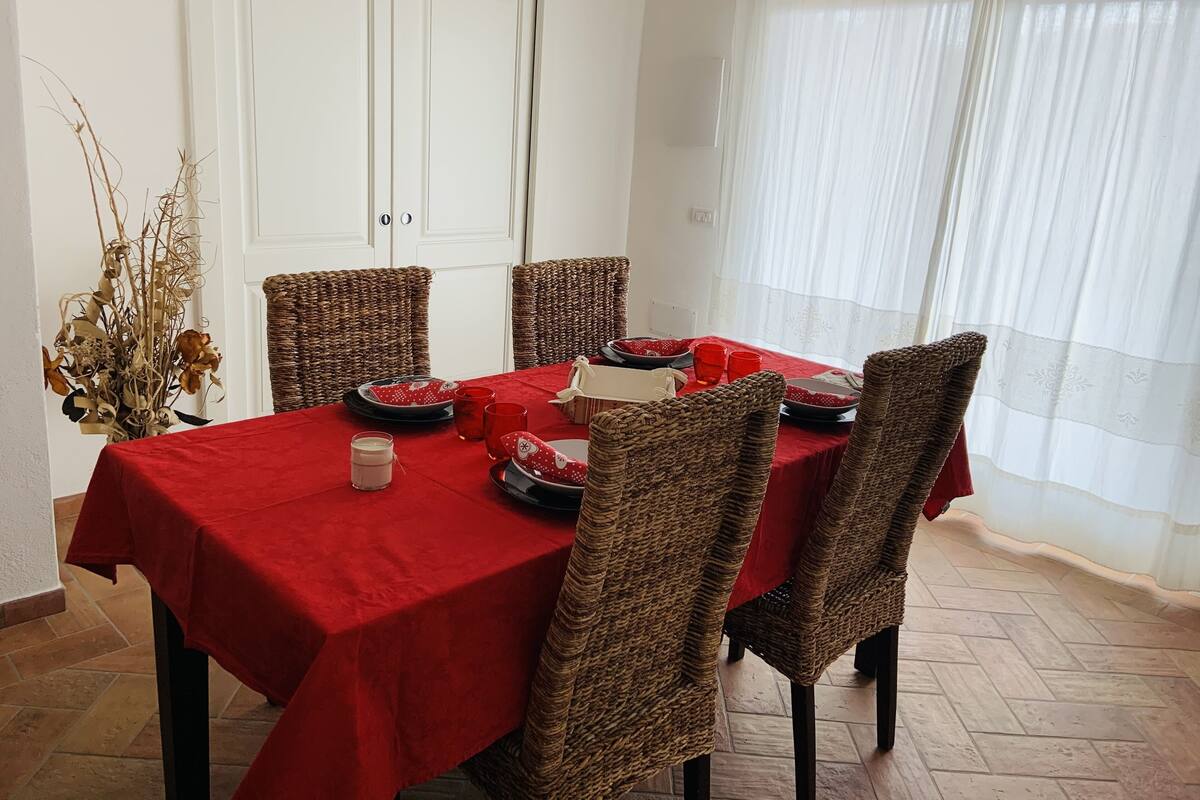 A dining area with a red tablecloth, wicker chairs, and a vase of dried flowers.