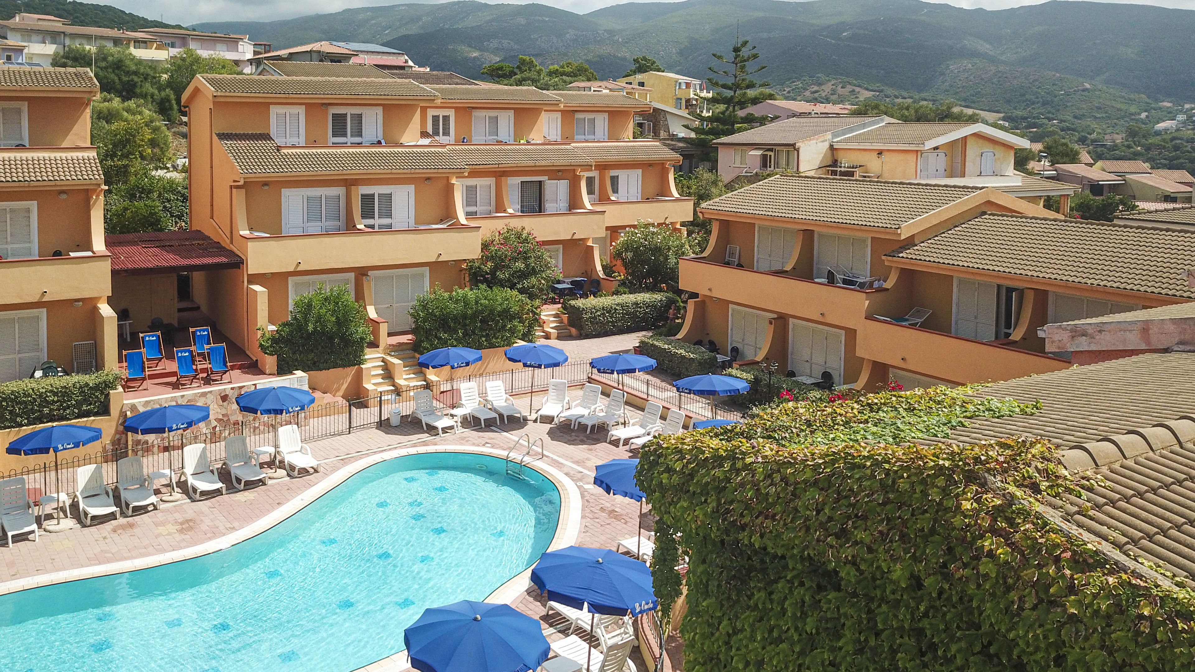 Uma área de piscina com espreguiçadeiras e guarda-sóis, cercada por edifícios com varandas e um cenário montanhoso.