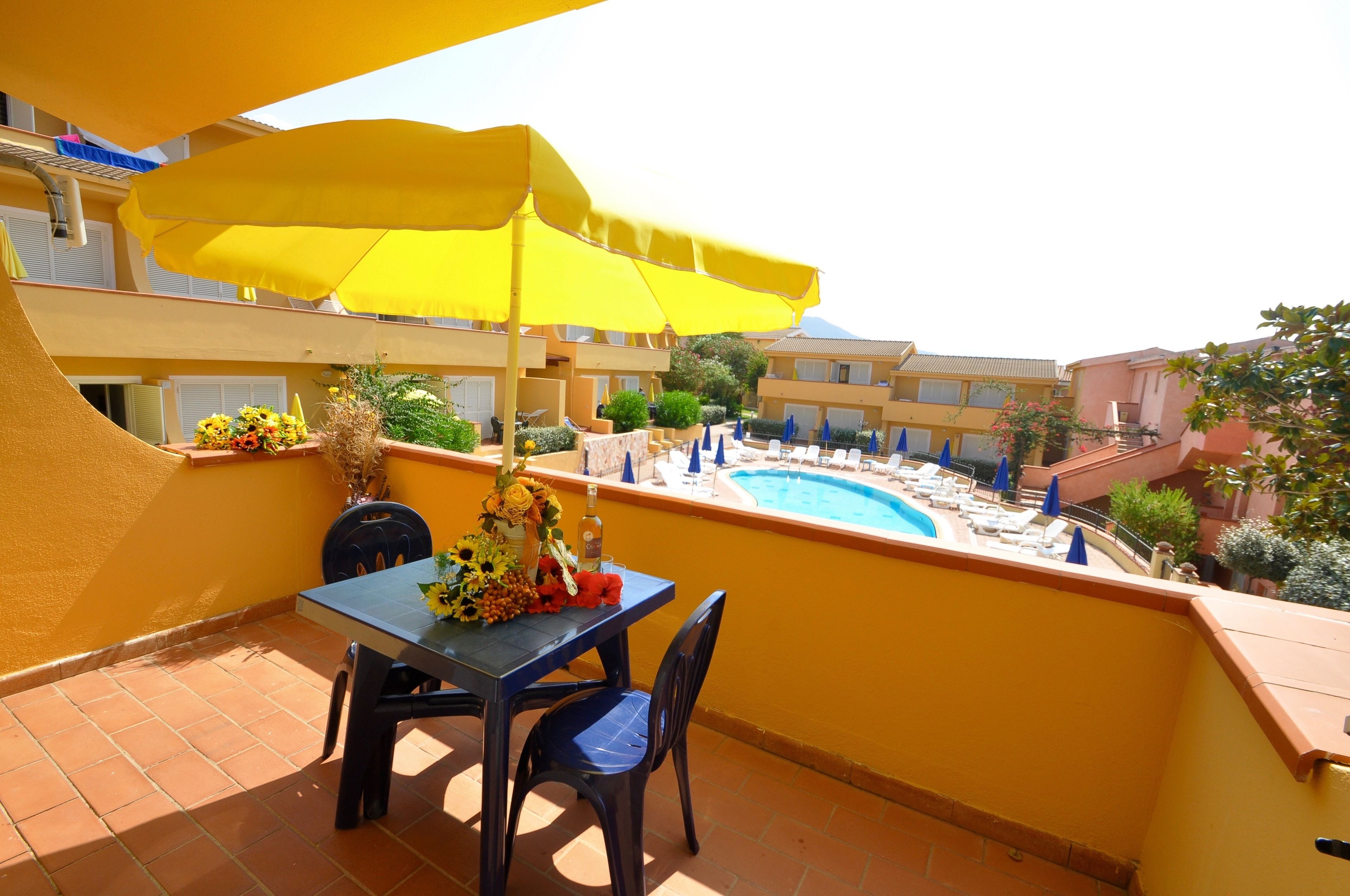 Een balkon met een gele parasol, een tafel met stoelen en een zwembad op de achtergrond.