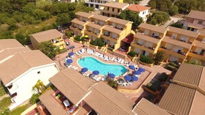 Ein Hotel mit Schwimmbad, umgeben von Liegestühlen und Sonnenschirmen, mehreren Balkonen und einer hügeligen Landschaft im Hintergrund.