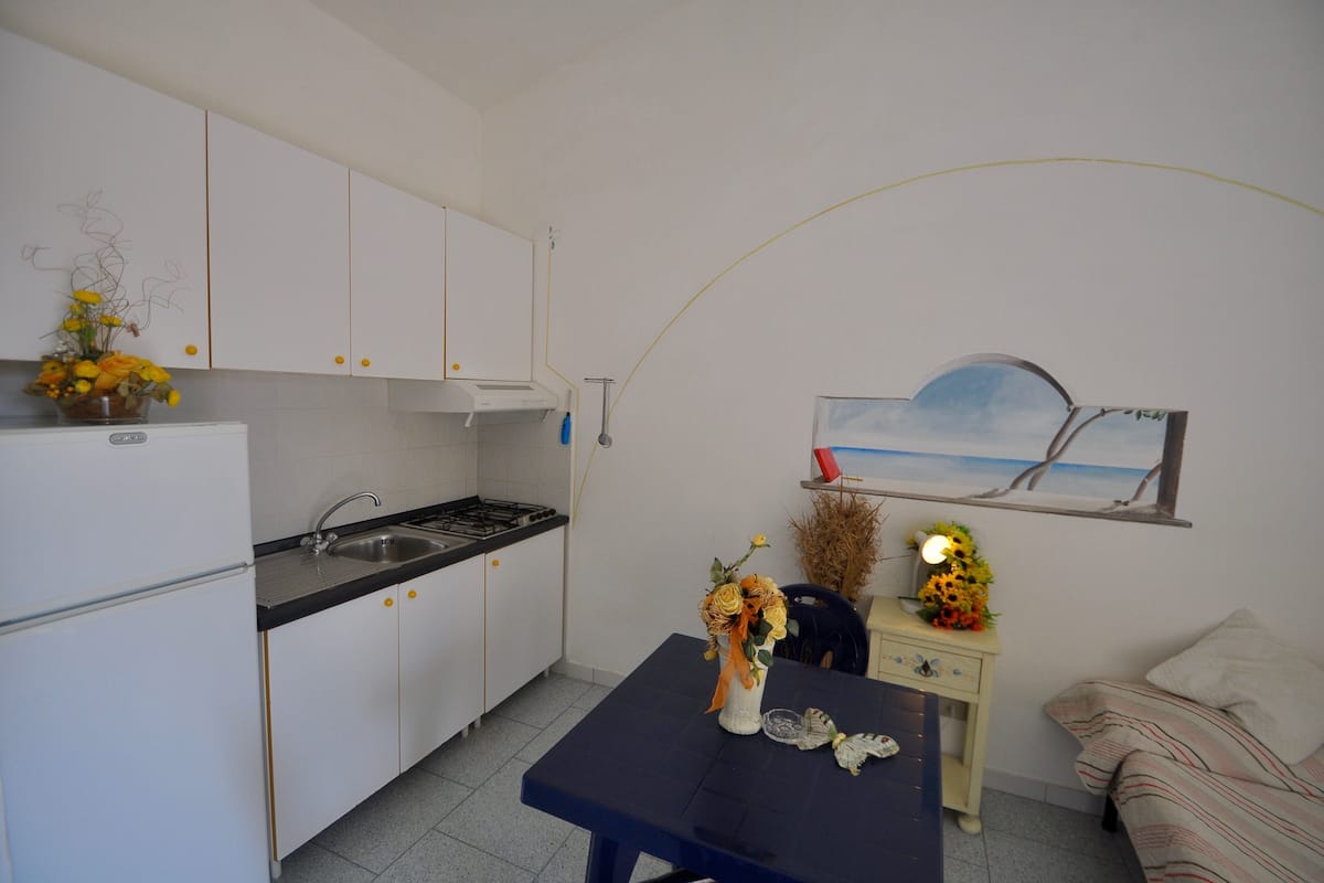 A compact kitchen with white cabinets, a black countertop, and a dining area with a table and chairs. A painting of a beach scene is on the wall.