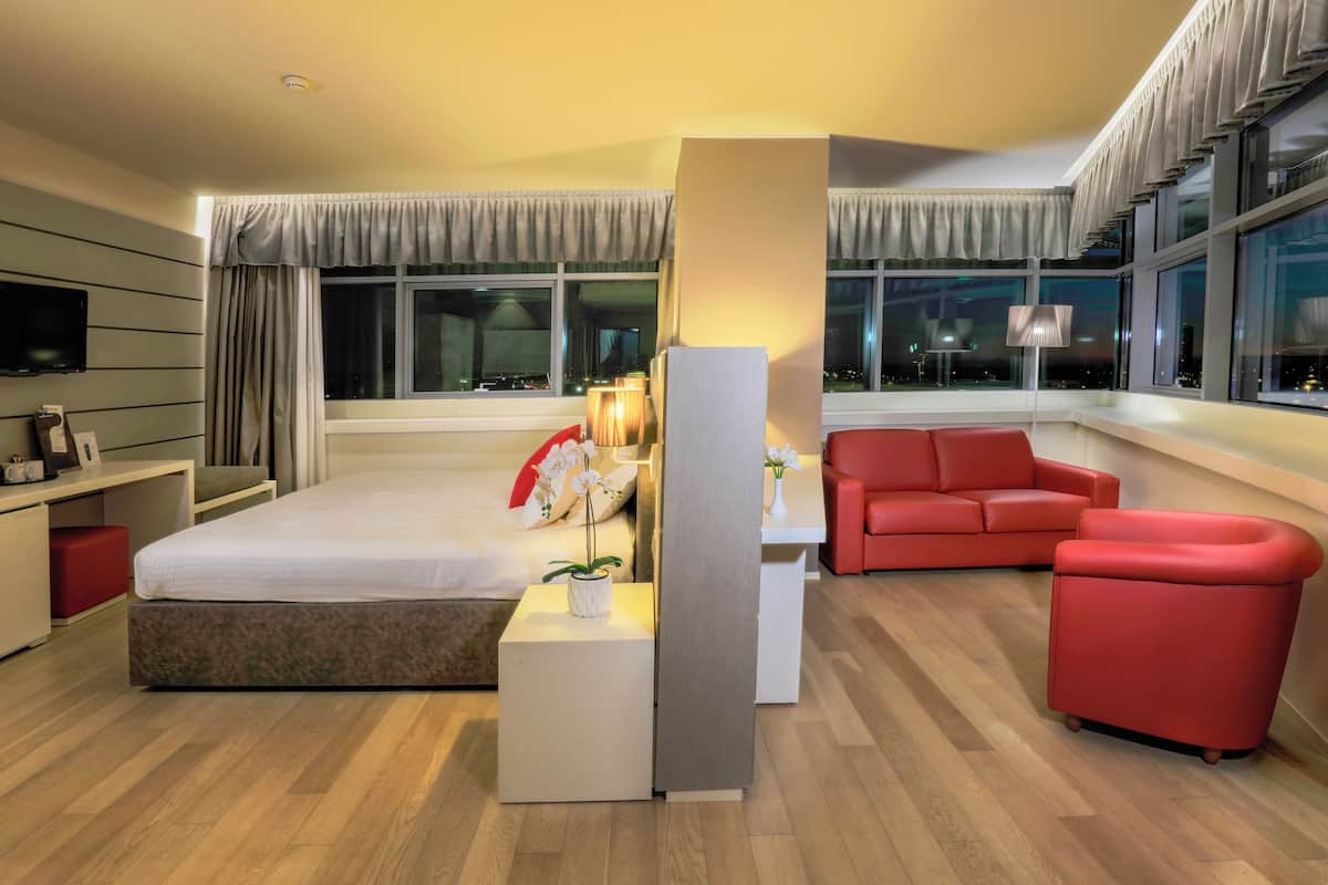 Una habitación de hotel moderna con una cama, un sofá rojo, una mesita de noche con una lámpara y un televisor en la pared.