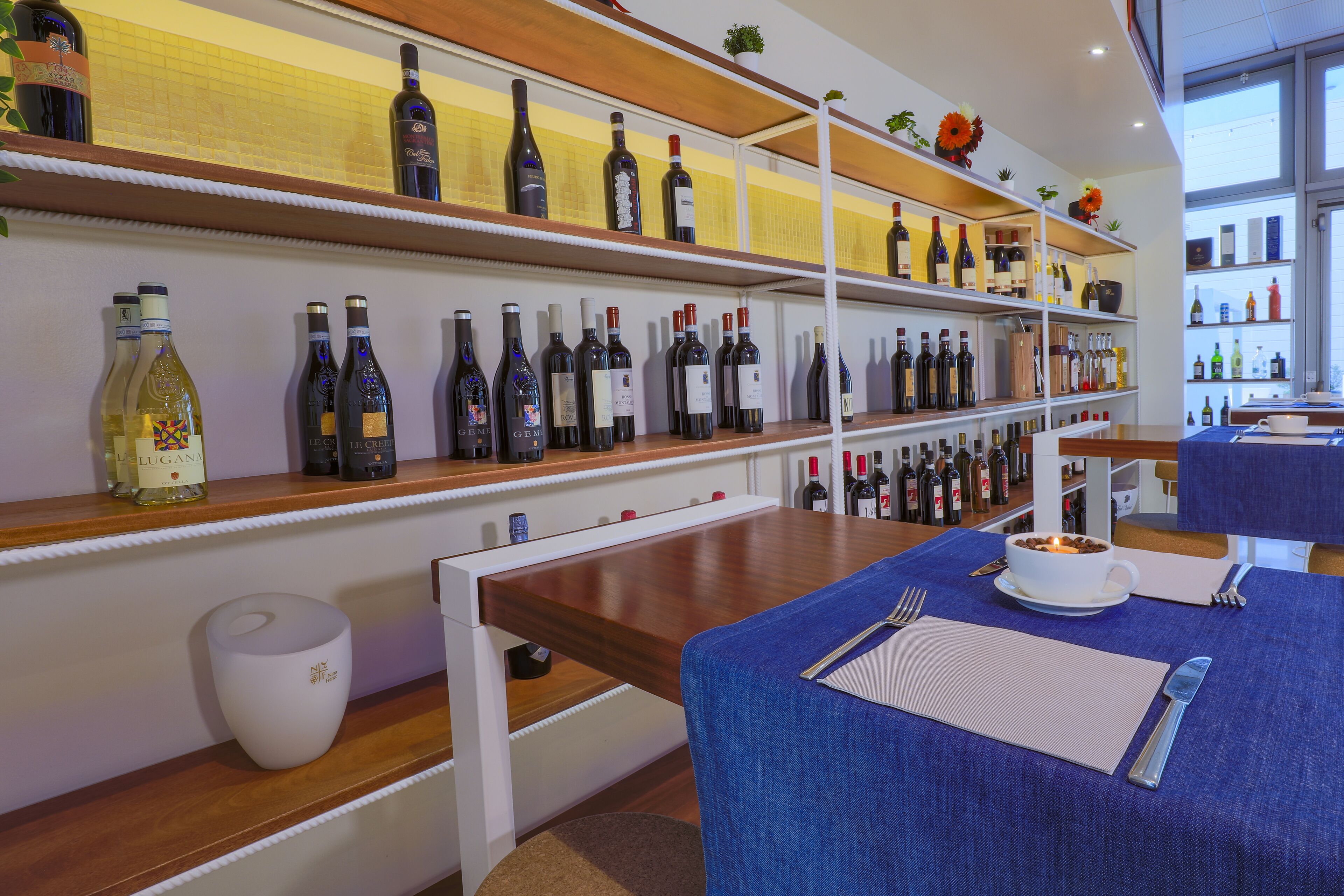 L’intérieur d’un restaurant, avec des bouteilles de vin exposées sur des étagères, une table à manger recouverte d’une nappe bleue et une bougie posée sur la table.