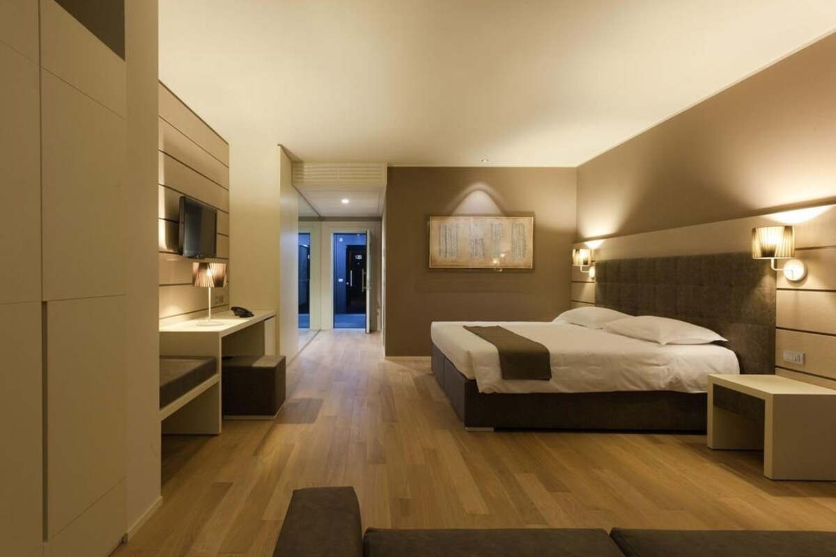 A modern hotel room with a large bed, bedside tables, a TV, and a view into another room.
