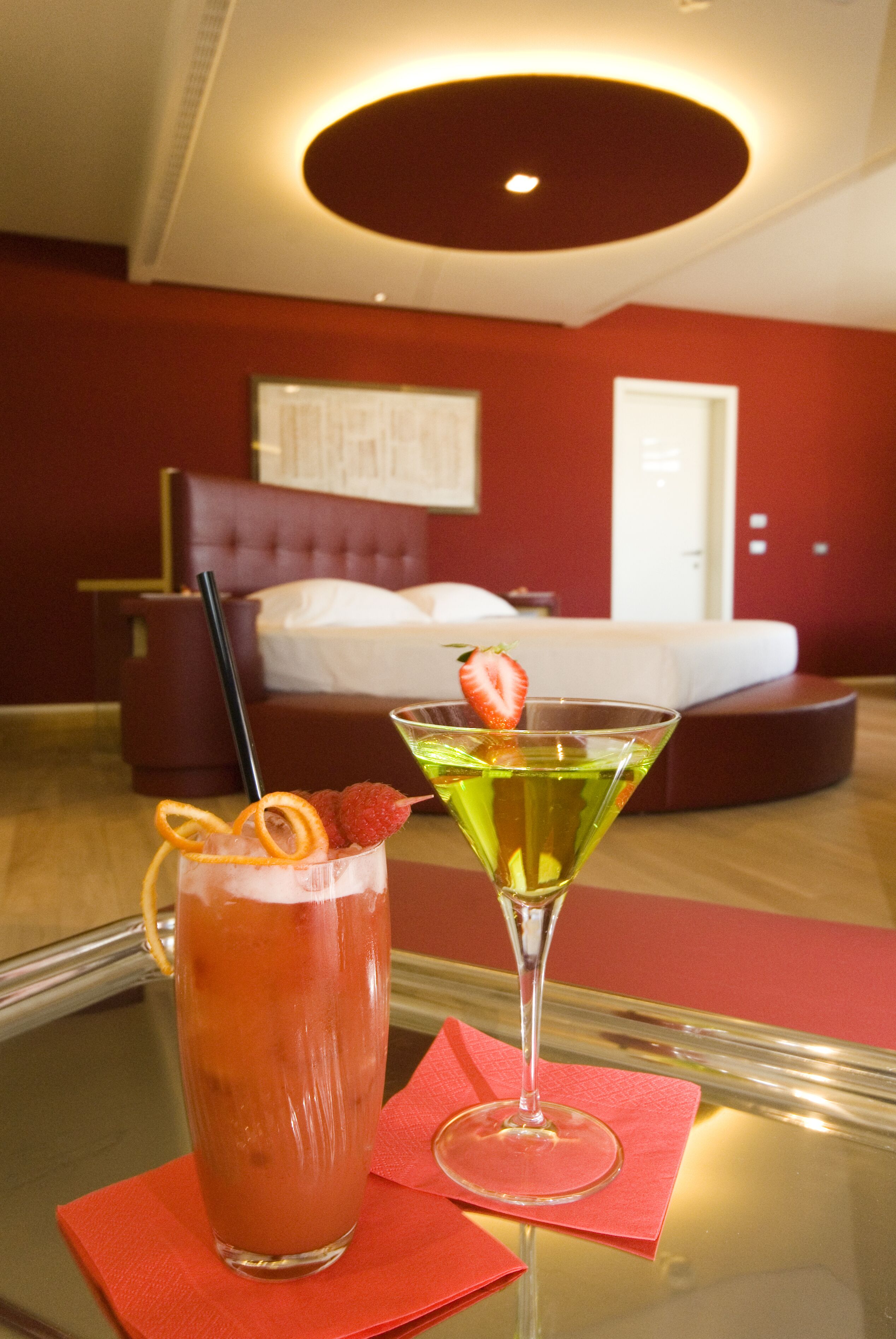 A room with a bed, a glass of red drink with a strawberry, and a martini glass with a green drink.