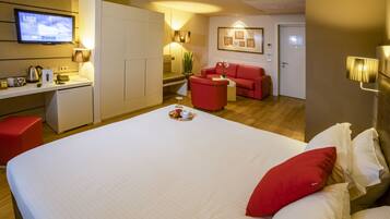 Quarto de hotel com uma cama grande, um sofá vermelho, uma escrivaninha com uma televisão e uma kitchenette pequena.