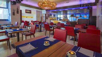 A restaurant with red chairs, wooden tables, and a bar area in the background.