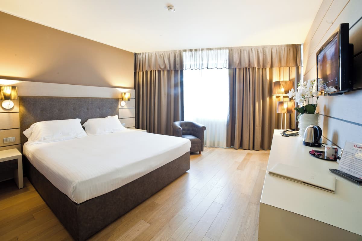 Una habitación de hotel con una cama grande, mesitas de noche, un escritorio con una computadora, una silla y cortinas.