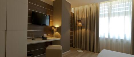 A hotel room with a bed, a desk with a TV, a lamp, and a window with curtains.