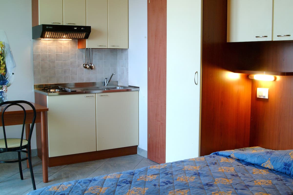 A compact room with a kitchenette, a bed with a blue patterned bedspread, and a wooden chair.