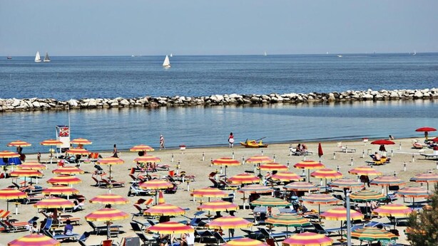 Una spiaggia con numerosi ombrelloni colorati e lettini, un molo roccioso e vele a distanza.