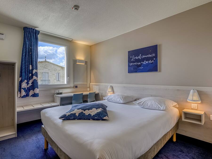 A hotel room with a bed, bedside tables, a window with a view, a built-in bench, and a wall-mounted artwork.