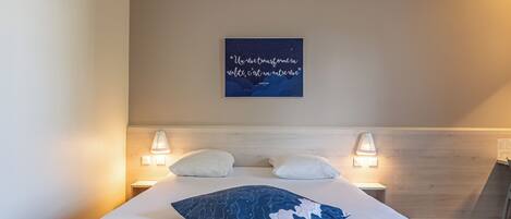 Habitación de hotel con cama, dos lámparas de noche, una luz en la pared y una cita decorativa.
