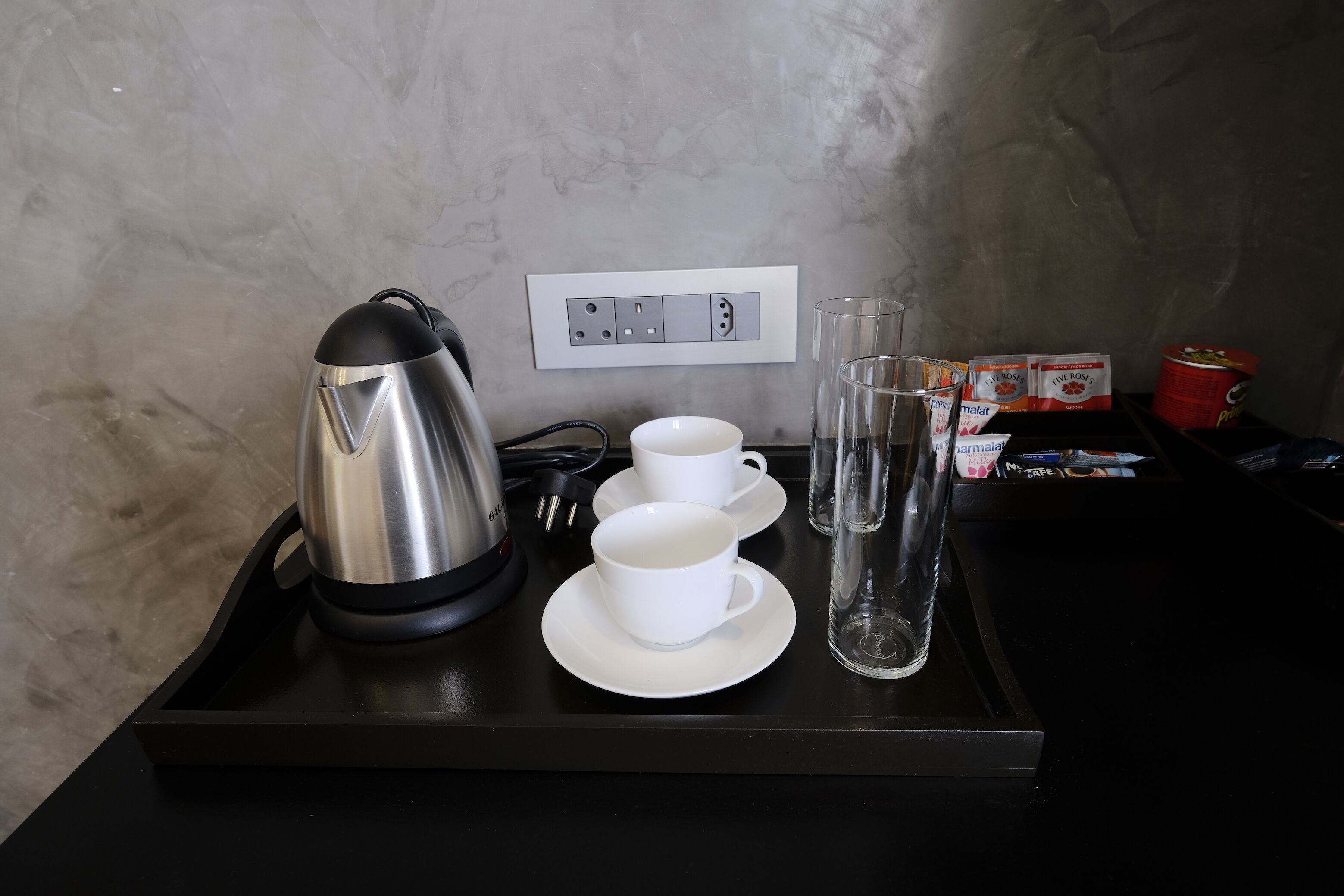 Una bandeja negra con una tetera, dos tazas blancas, un vaso y algunos alimentos envasados sobre una mesa frente a una pared con textura.