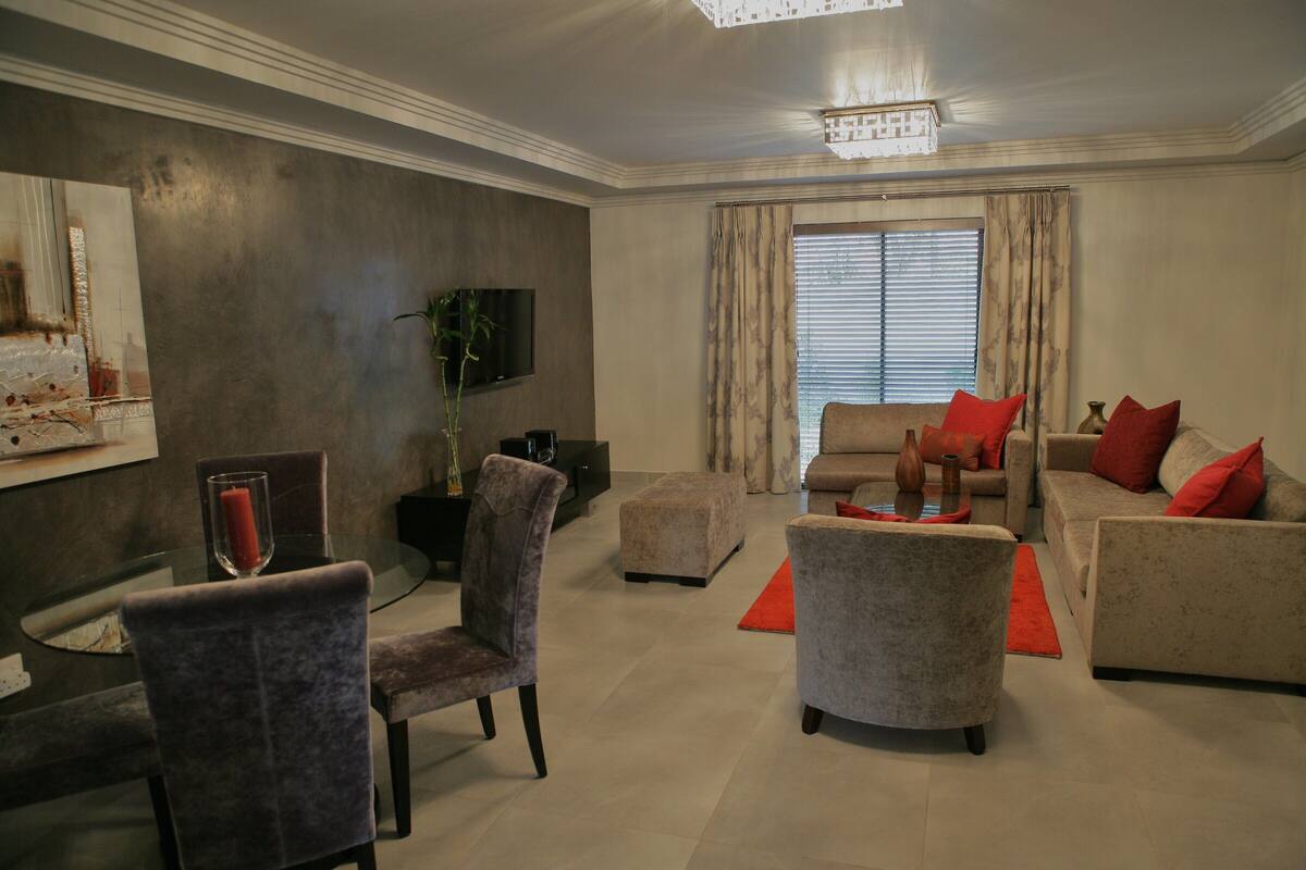 Un salon moderne avec une table basse en verre, deux fauteuils, un canapé et un téléviseur fixé au mur.
