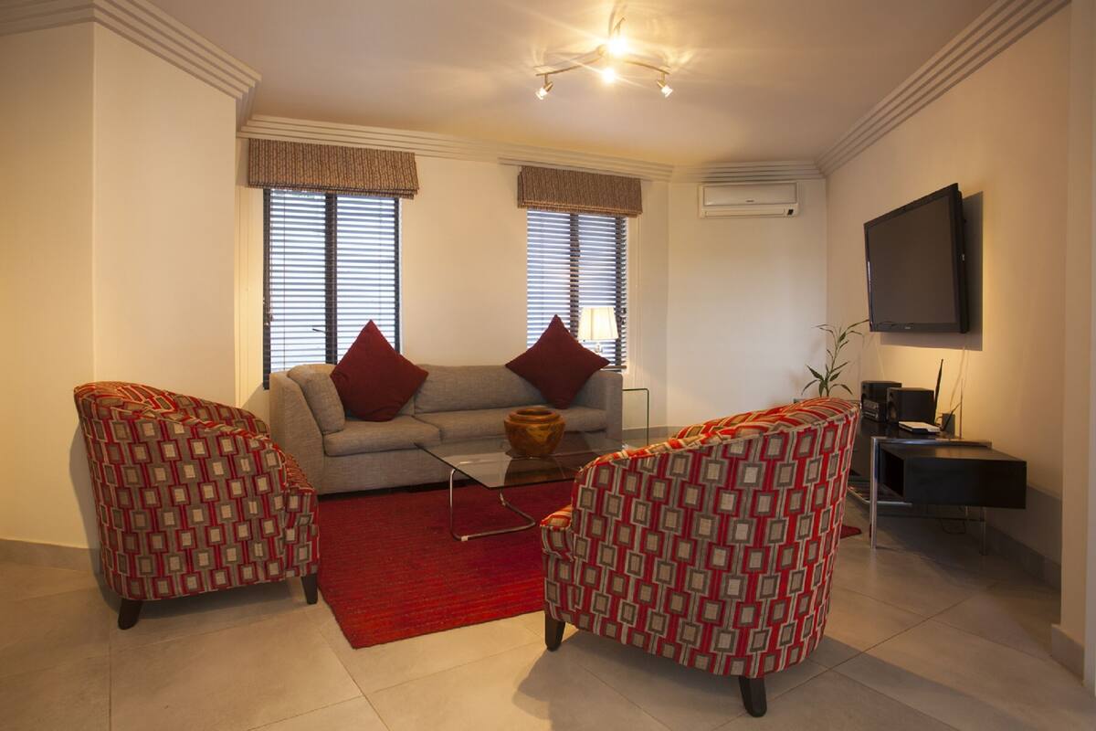 Un salon avec un canapé, deux fauteuils à motifs, un tapis rouge et un téléviseur fixé au mur.