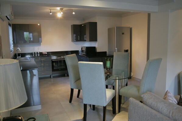 A modern kitchen with dark cabinets, a stainless steel refrigerator, and a dining area with a glass table and two chairs.