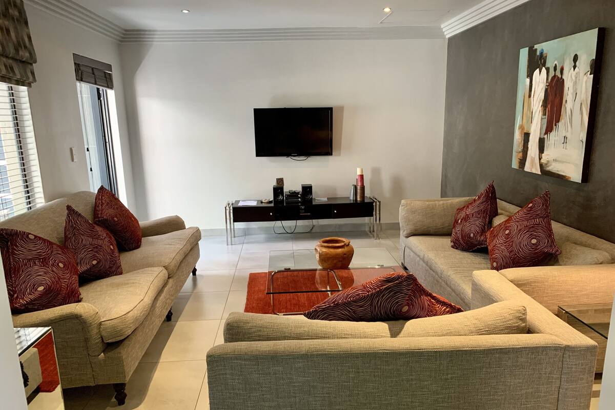 Un salon moderne avec deux canapés beige, des coussins à motifs rouges, une table basse en verre, un téléviseur à écran plat fixé au mur et une console en bois foncé sur laquelle sont posés des appareils électroniques.