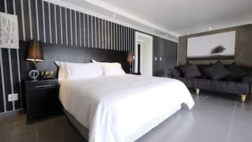 A modern bedroom with a large bed, a dark headboard, a nightstand with a lamp, a black sofa, and a large framed picture on the wall.