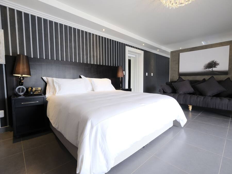 A modern bedroom with a large bed, a dark headboard, a nightstand with a lamp, a black sofa, and a large framed picture on the wall.