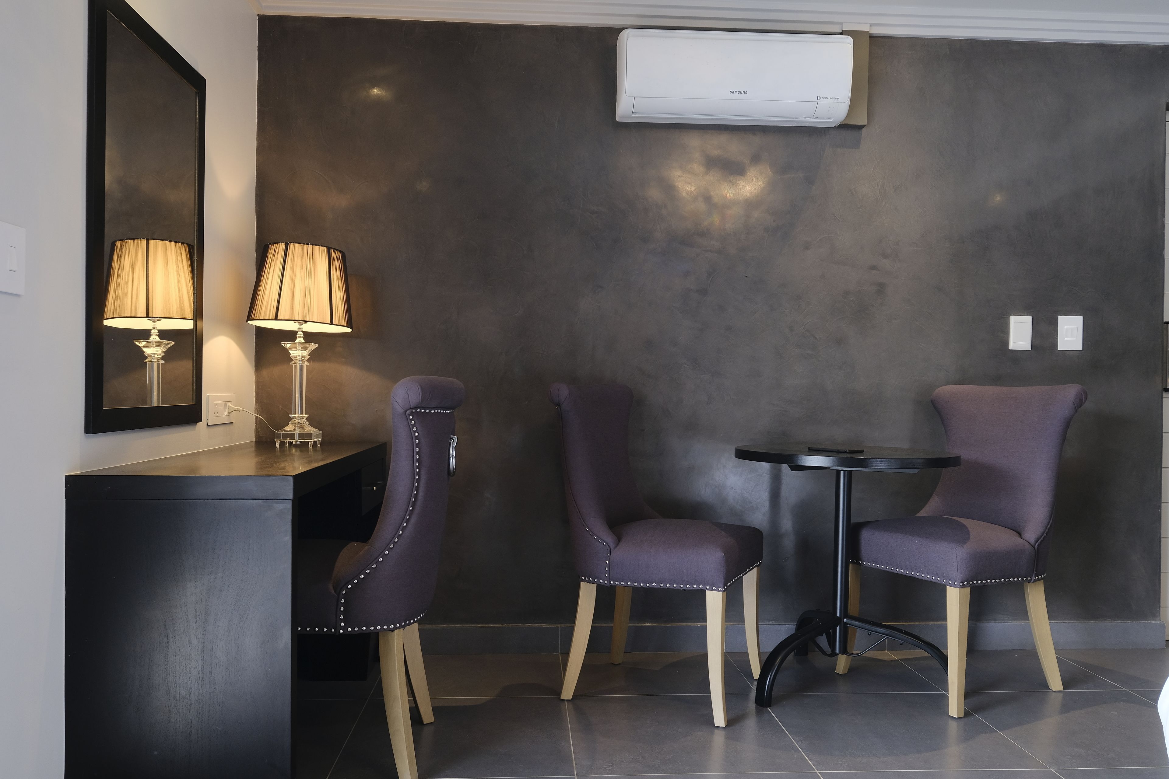 Een moderne kamer met een donkere, structuurrijke muur, een ronde tafel, twee paarse stoelen, een spiegel en een lamp.