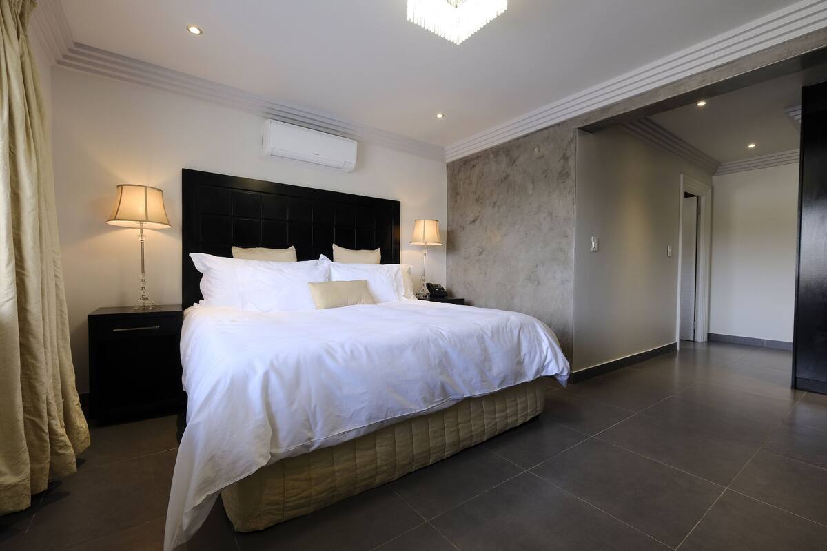 Une chambre d’hôtel moderne avec un grand lit, deux tables de chevet avec des lampes, une tête de lit sombre et un grand miroir.