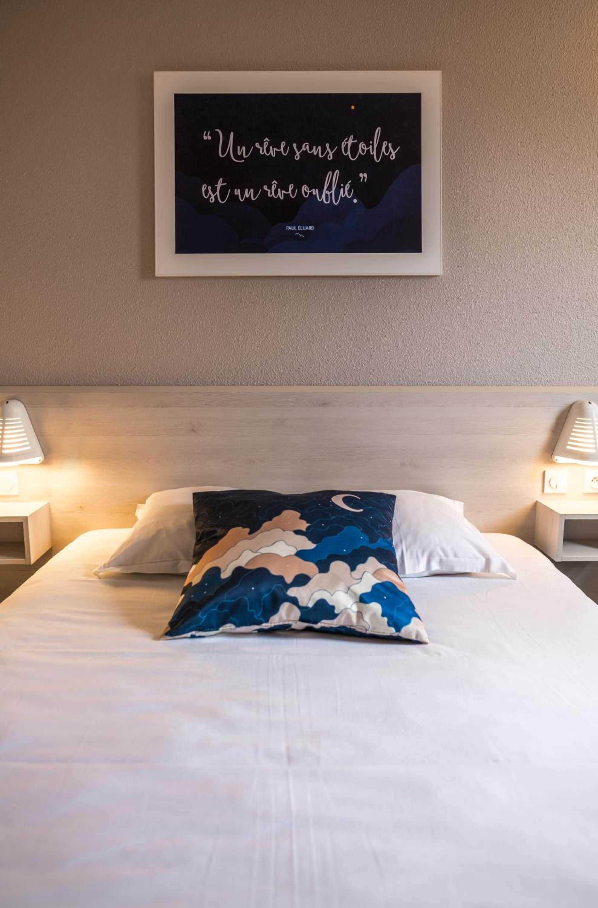 Cama bem arrumada com travesseiro padronado em branco e azul, uma citação emoldurada na parede e duas luminárias de cabeceira.