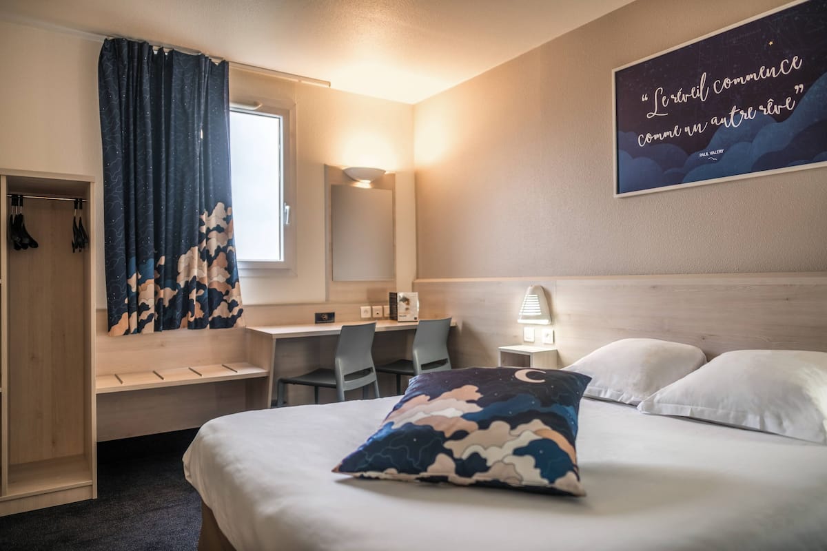 A hotel room with a bed, a desk, a chair, a wardrobe, and a wall with a quote.