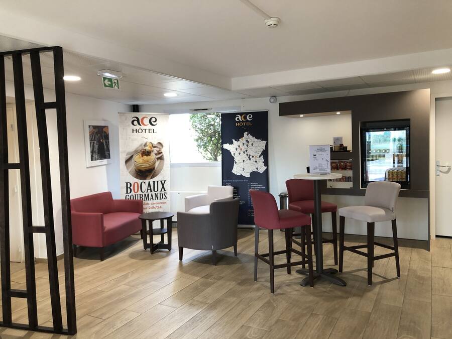 Ein Hotel-Empfangsbereich mit einer Karte Frankreichs, einem Schild für 'ace HOTEL' und einem Plakat mit der Aufschrift 'BOCAUX GOURMANDS'. Es sind rote und graue Stühle sowie ein kleiner Tisch vorhanden.