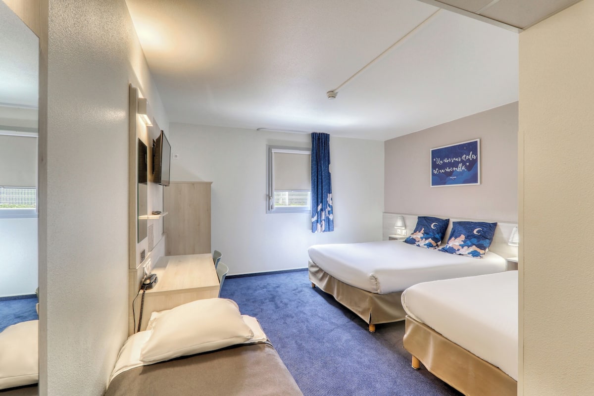 Une chambre d’hôtel avec deux lits, un bureau, une chaise, une fenêtre avec des rideaux et un téléviseur fixé au mur.