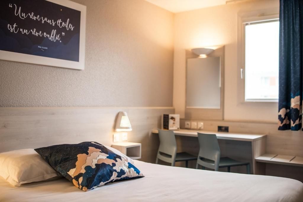 A hotel room with a bed, a desk, a chair, a lamp, and a wall-mounted mirror.