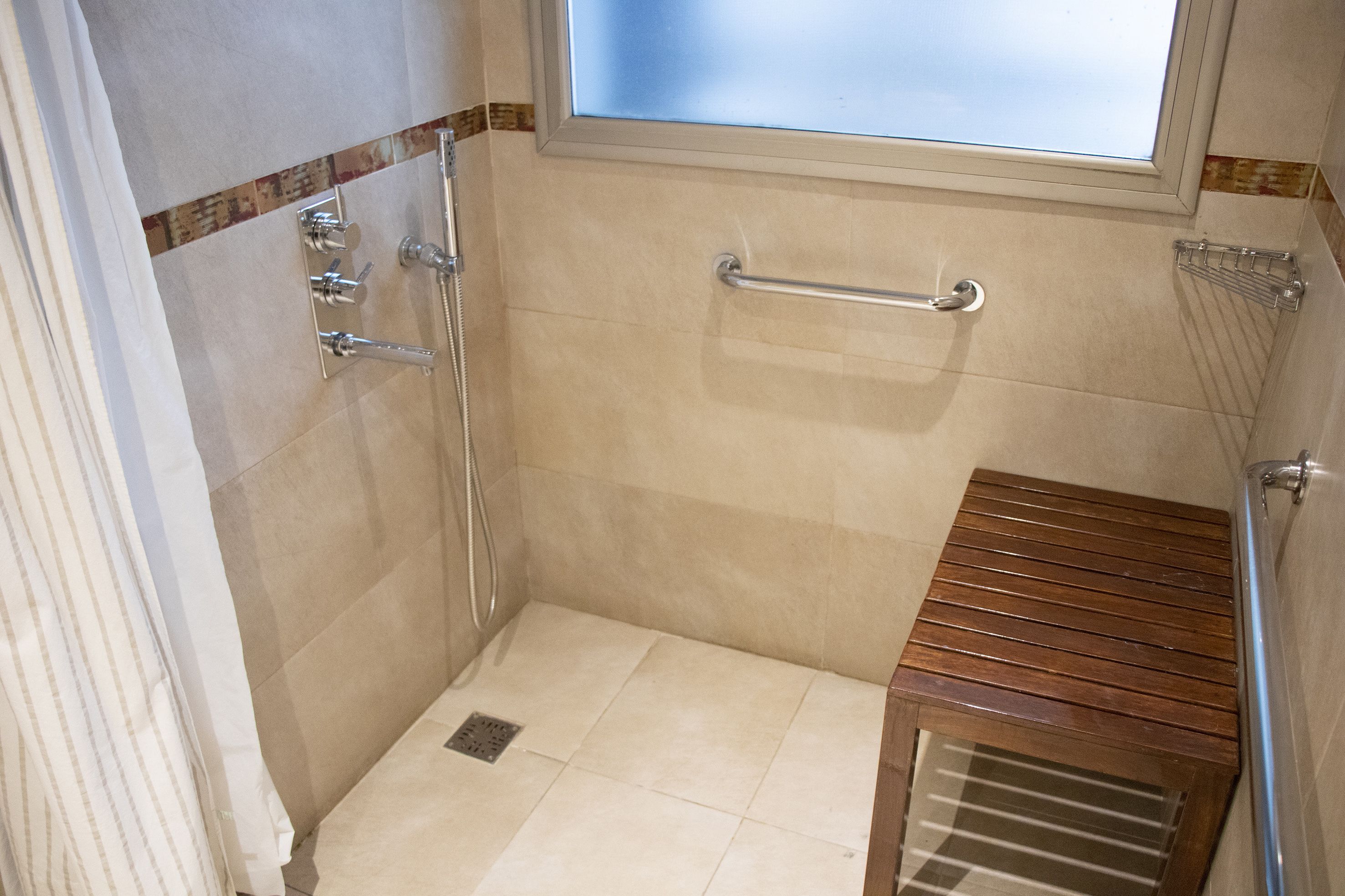 A small, tiled bathroom with a shower, a wooden bench, and a window.