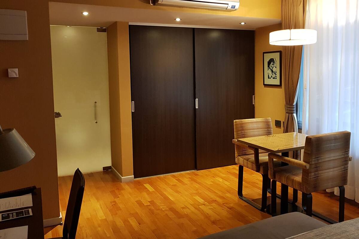 Una habitación de hotel con piso de madera, una mesa de comedor con sillas, una cama, un armario y un aire acondicionado instalado en el techo.