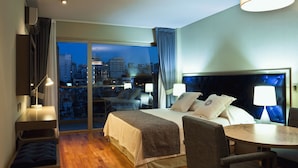 Habitación de hotel con cama, escritorio, silla, mesa y vistas a la ciudad al anochecer.