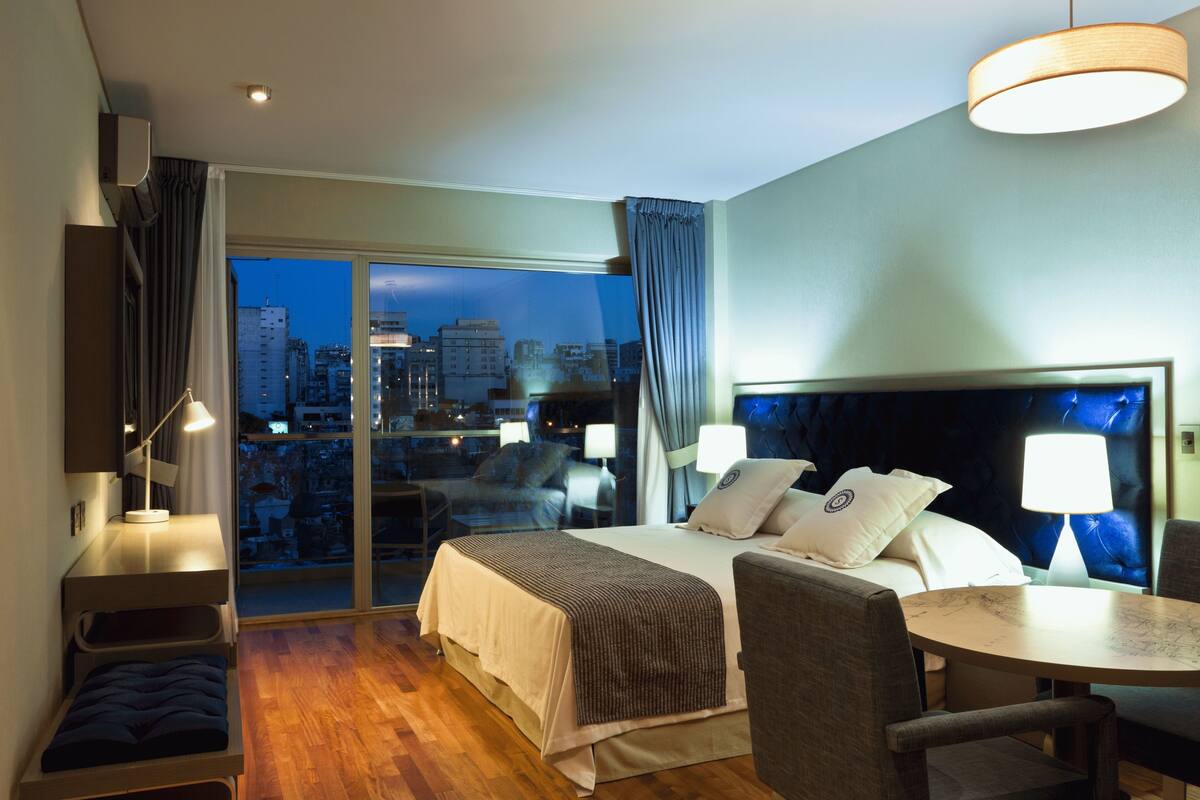 Habitación de hotel con cama, escritorio, silla, mesa y vistas a la ciudad al anochecer.