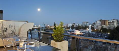 A rooftop terrace with metal chairs and tables, a potted plant, and a cityscape view at dusk.