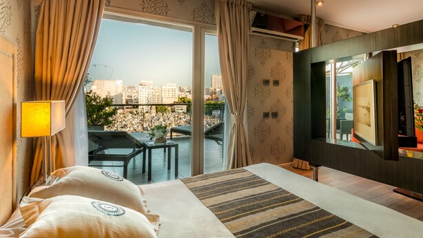 Habitación de hotel con una cama grande, televisión, balcón con vistas a la ciudad y una pequeña mesa con una planta.