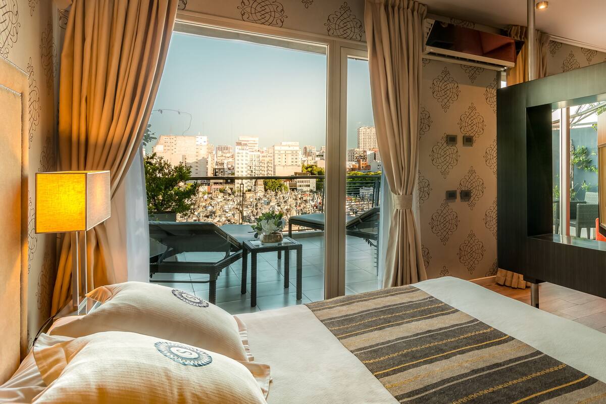 Habitación de hotel con una cama grande, televisión, balcón con vista a la ciudad y una mesa con sillas.