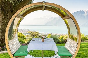 Uma estrutura circular de madeira com uma mesa e bancos, com vista para um lago e montanhas.