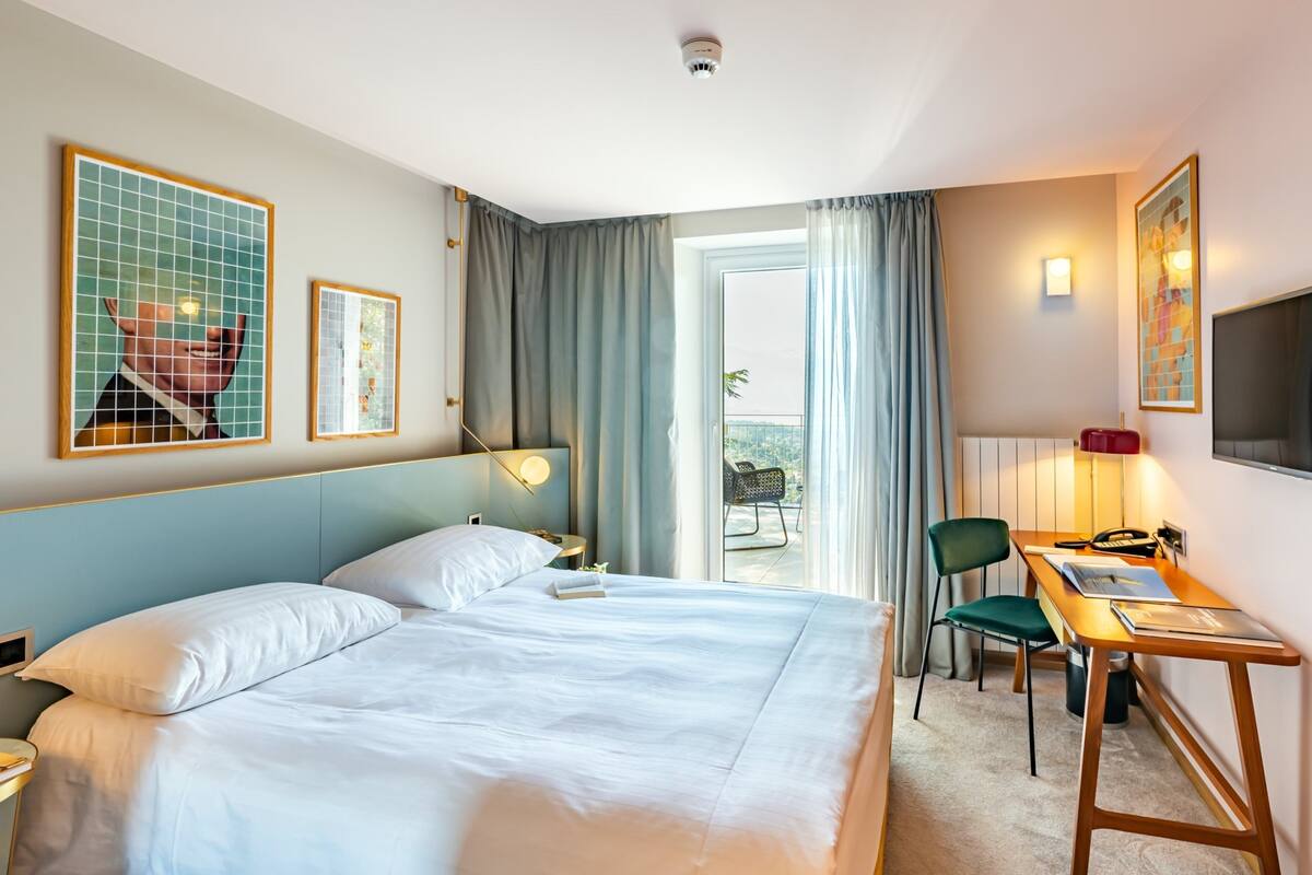 A hotel room with a large bed, a desk, a chair, and a balcony with a view.