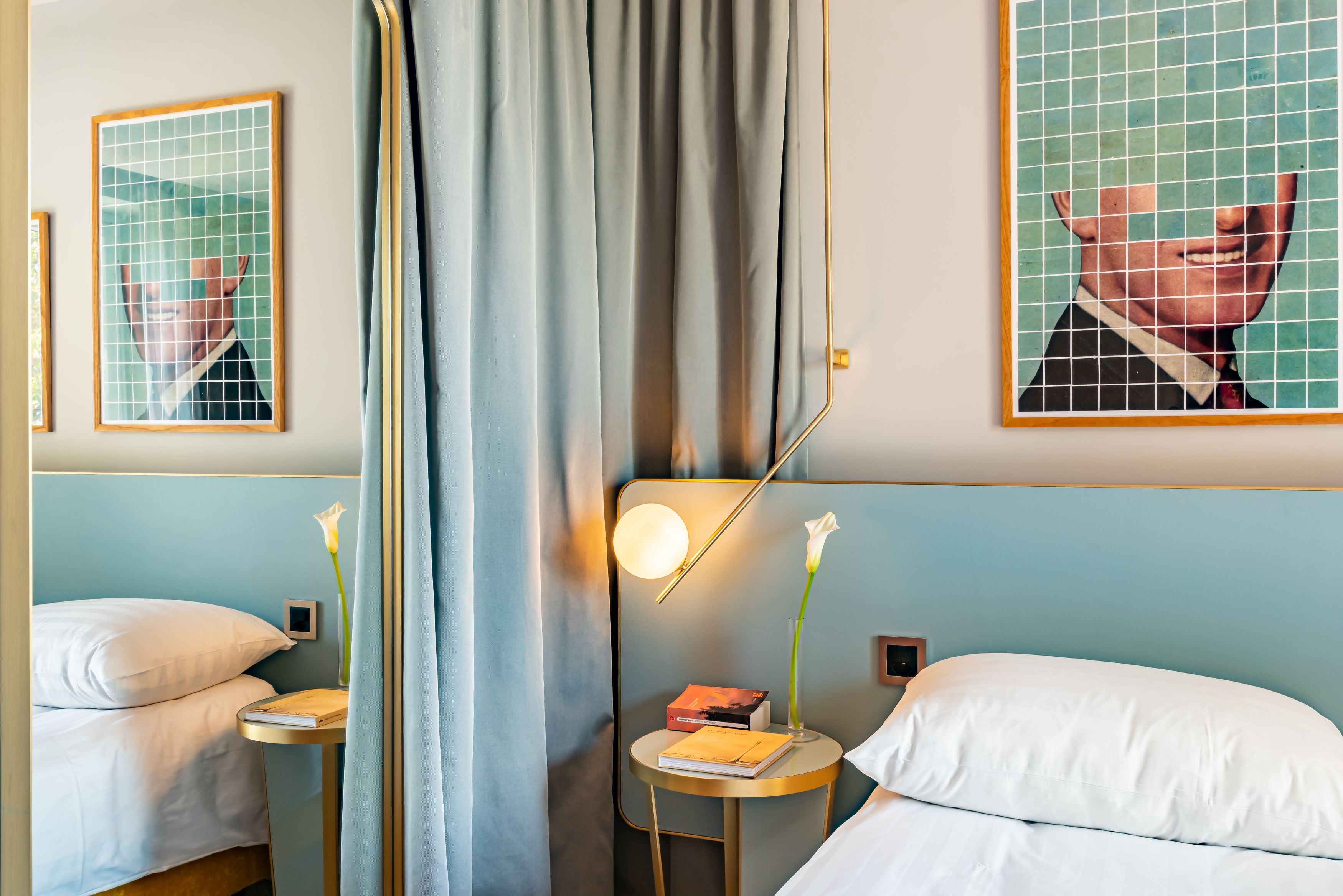 A hotel room with two beds, a window with curtains, a bedside table with a book and a vase of flowers, and a wall-mounted mirror.
